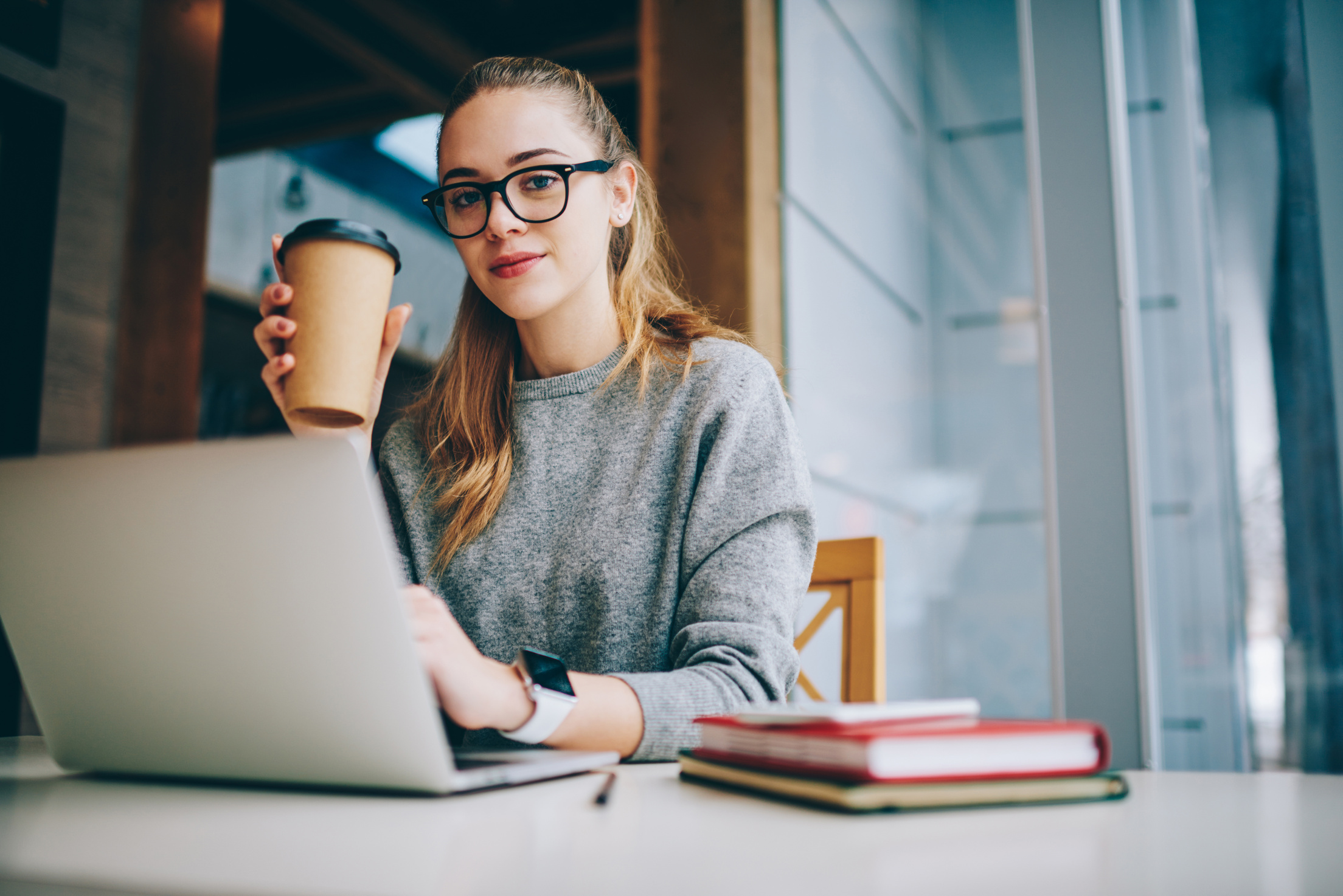 Portrait of pretty female copywriter enjoying morning coffee during break at favourite cafeteria, attractive software developer looking at camera after working on laptop computer connected to wifi